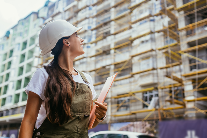 una mujer con un casco de obra y unos planos frente a un edificio en construcción